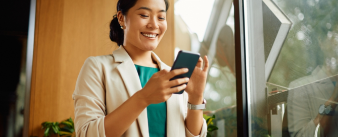 Young professional woman smiling at her phone in a warm, sunlit office space with greenery in the background