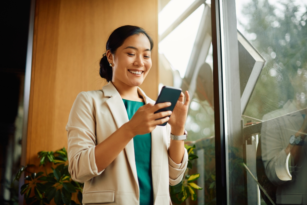 Happy,Asian,Businesswoman,Text,Messaging,On,Cell,Phone,While,Standing Young professional woman smiling at her phone in a warm, sunlit office space with greenery in the background